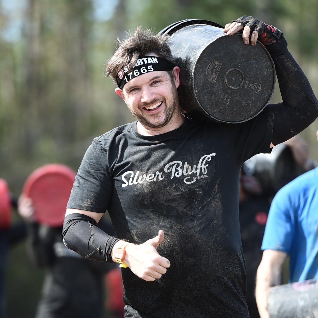 Jay Dolan in a Spartan race carrying a bucket while smiling and giving a thumbs up